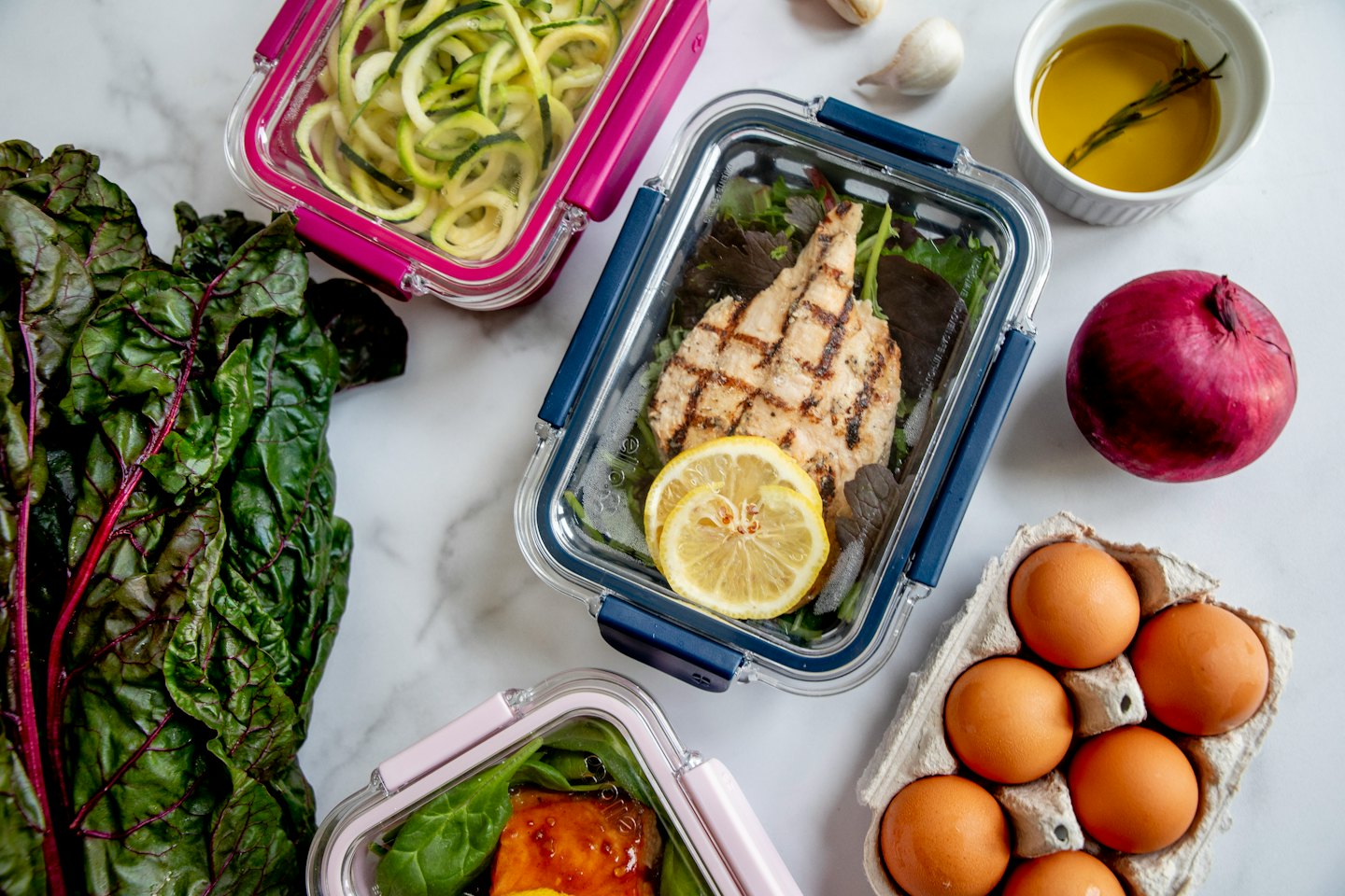 Three meal prep containers with healthy food on a white marble counter.